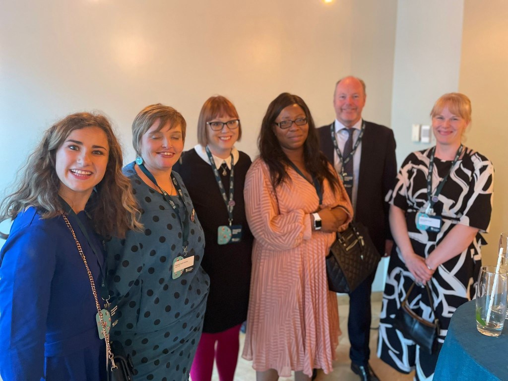 Standing in a line, from left, a lady in a blue dress with long wavy hair, Georgia Vine. A lady in a green spotty dress and short hair, Rachel Booth-Gardiner. A lady in a black dress with white collar and pink tights and short bob, Natalie Hicks, a Black lady in a pink long sleeve dress with long wavy black hair, Anne-Marie Fadare, a man in a suit, Steve Ford, a woman in a black and white knee length dress, blond hair put up, Sam Pywell.,
The women are Founding Members of AbleOTUK and Steve Ford is the Chair of the Royal College of Occupational Therapists